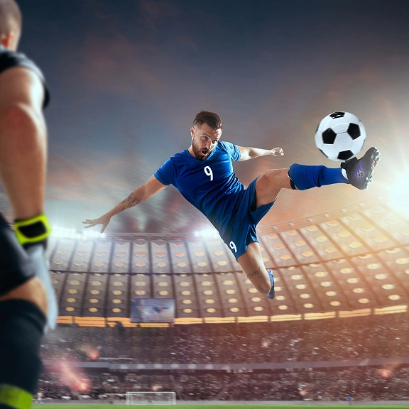 Piłkarz w niebieskim stroju wykonuje dynamiczny strzał na stadionie podczas meczu.